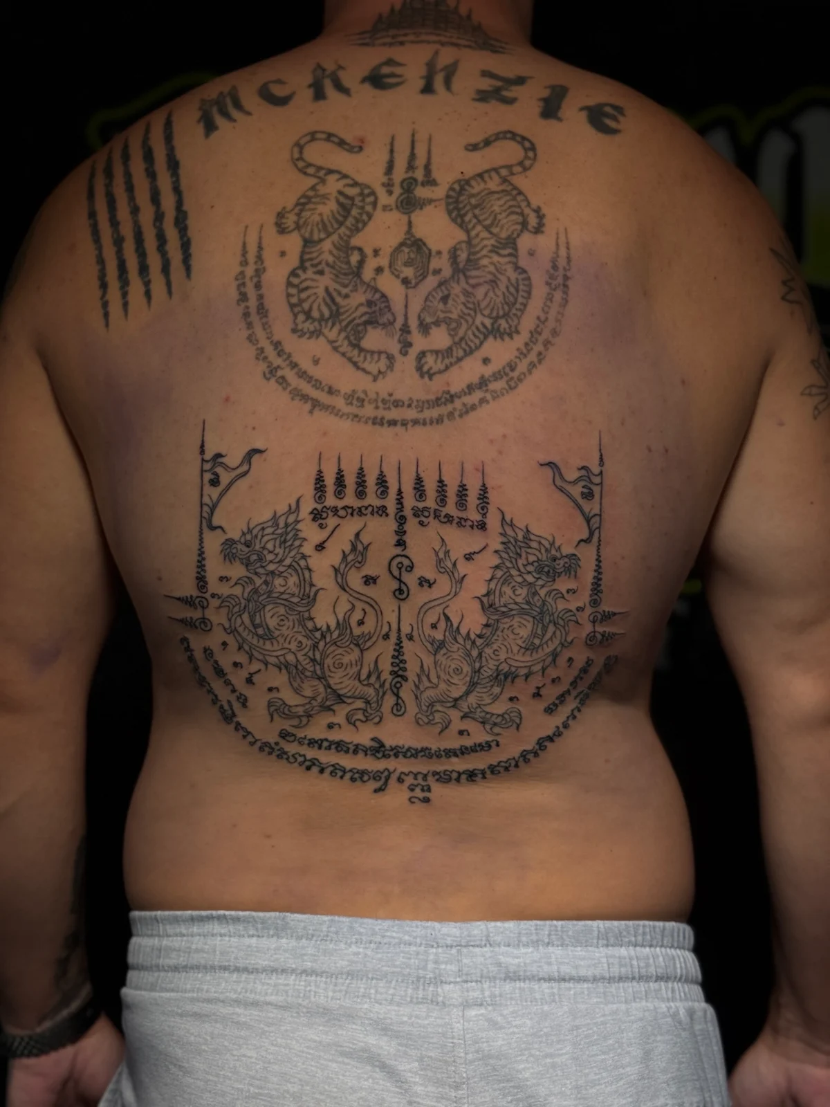 Large back Sak Yant with Suea Koo twin tigers, Yant Singha mythical lions, Hah Taew five lines, and MCKENZIE script lettering at Bloodline Tattoo Phuket.