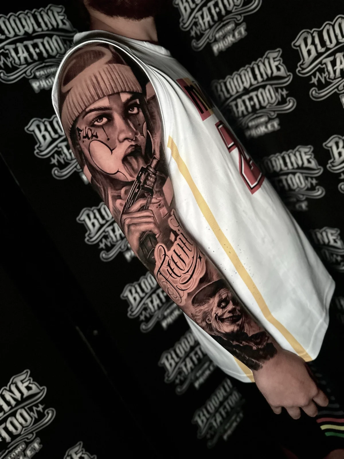 Chicano arm sleeve with payasa portrait, handgun, script lettering, and Joker face at Bloodline Tattoo Phuket.