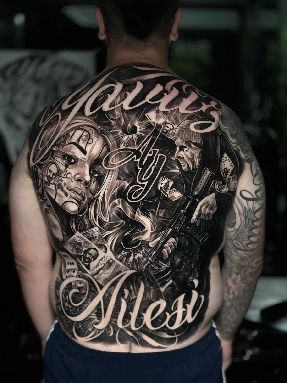 Chicano back tattoo with Yavuz Ailesi script, money, rifle portrait, and Australian banknotes at Bloodline Tattoo Phuket.