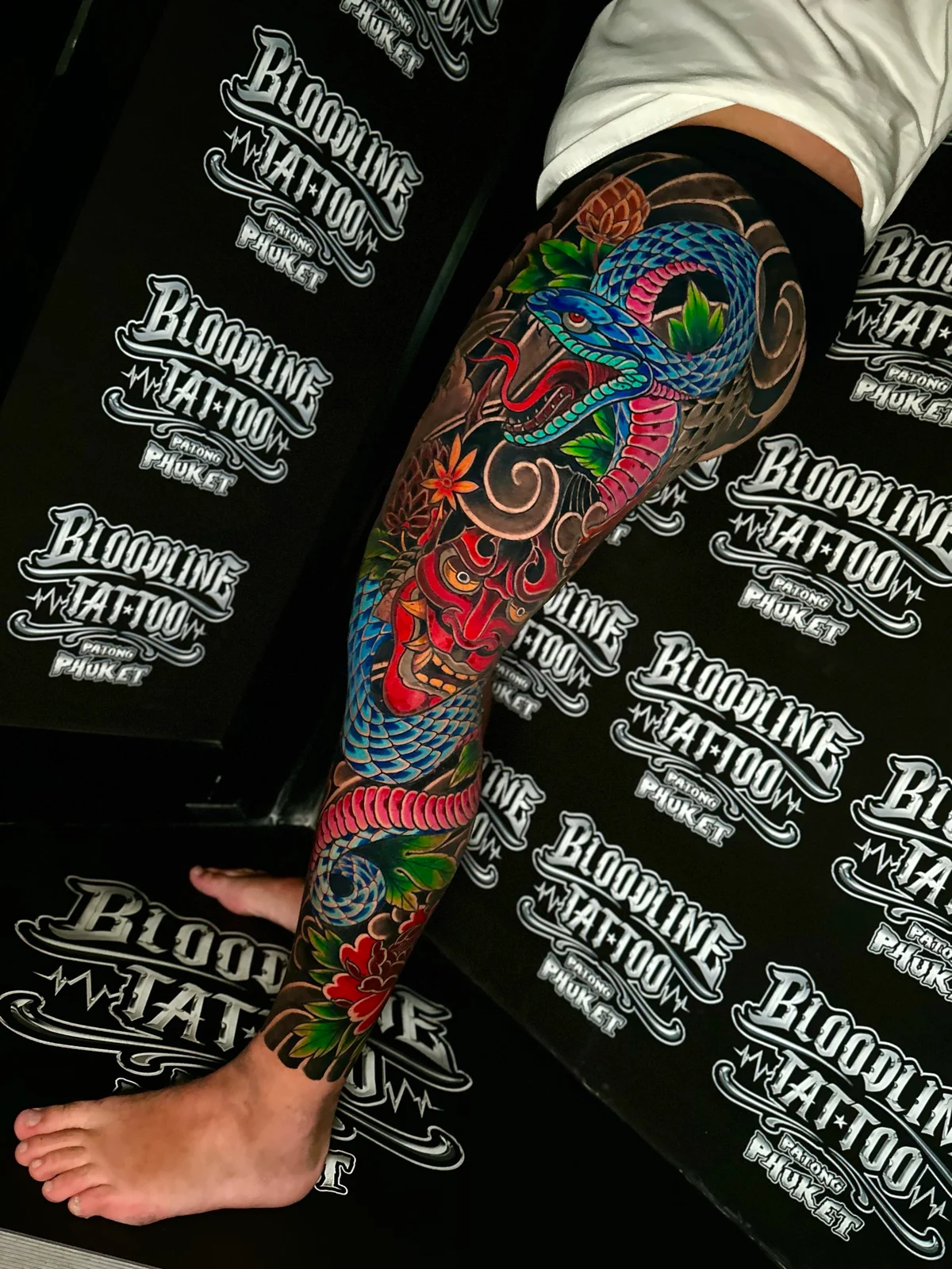 Japanese colour full-leg tattoo with a coiled blue and pink snake, a red Hannya mask, and floral accents in black wind-bar shading at Bloodline Tattoo Phuket.