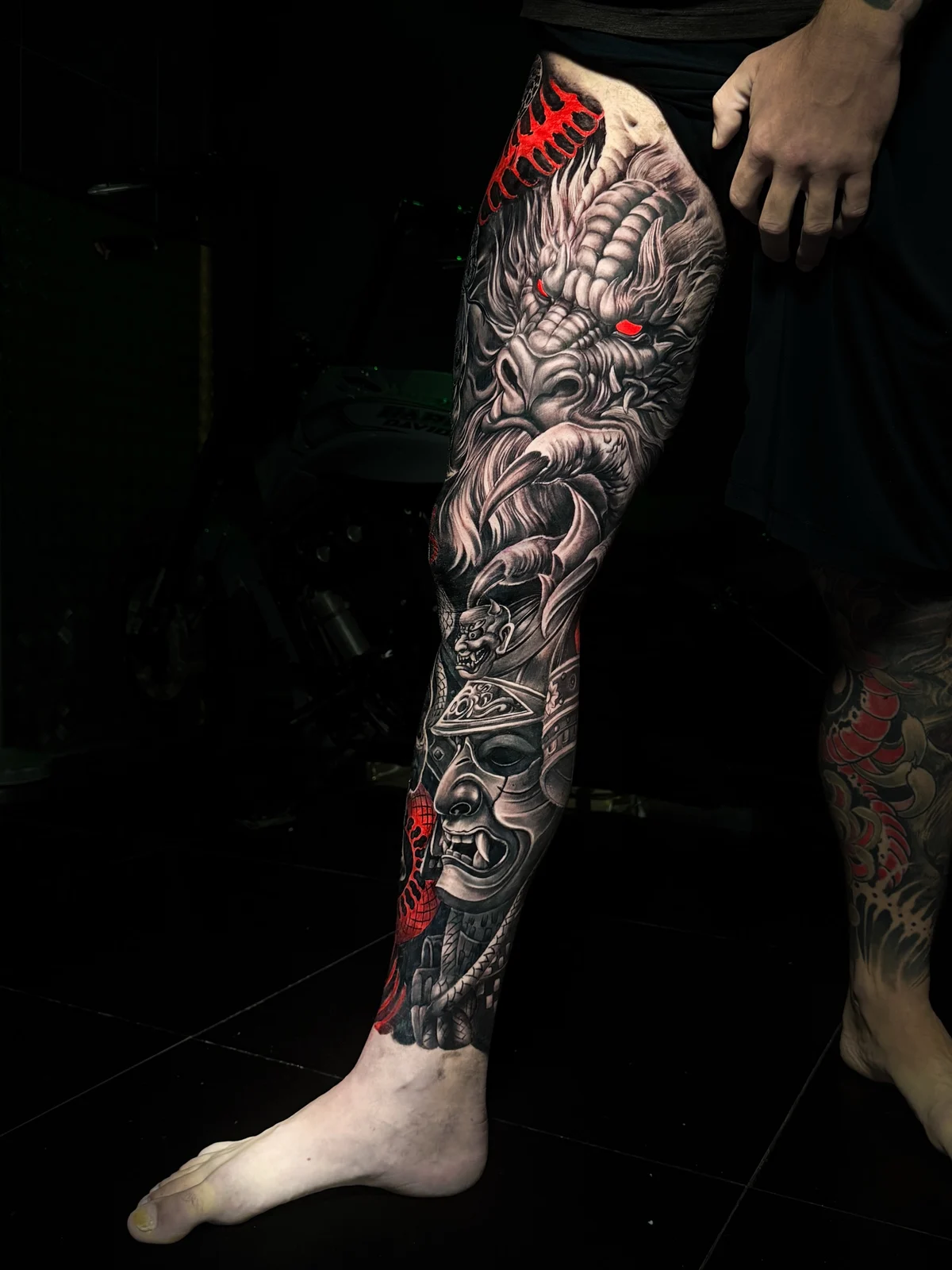 Japanese full leg sleeve with dragon head, glowing red eyes, samurai menpo, and oni mask in neo-irezumi black and grey at Bloodline Tattoo Phuket.