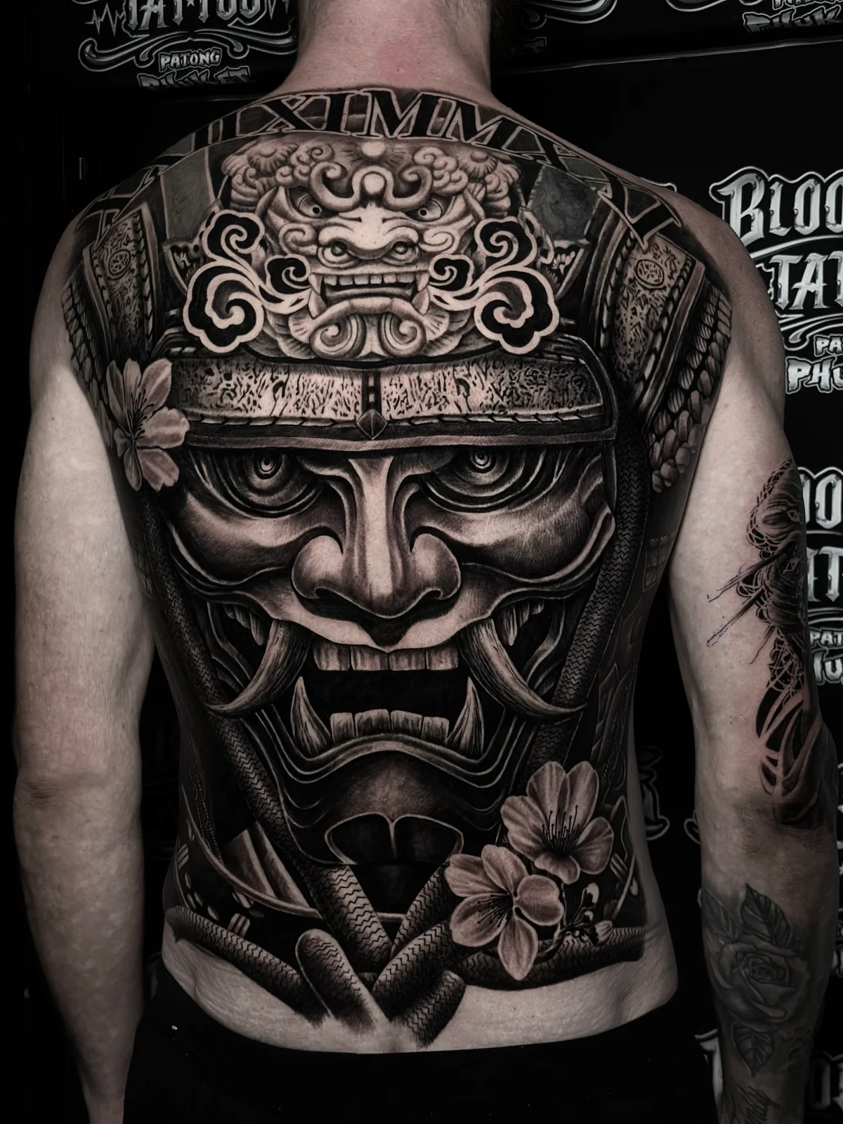 Japanese black and grey full-back samurai mask with shishi crest, cherry blossoms, and roman numerals at the neckline at Bloodline Tattoo Phuket.