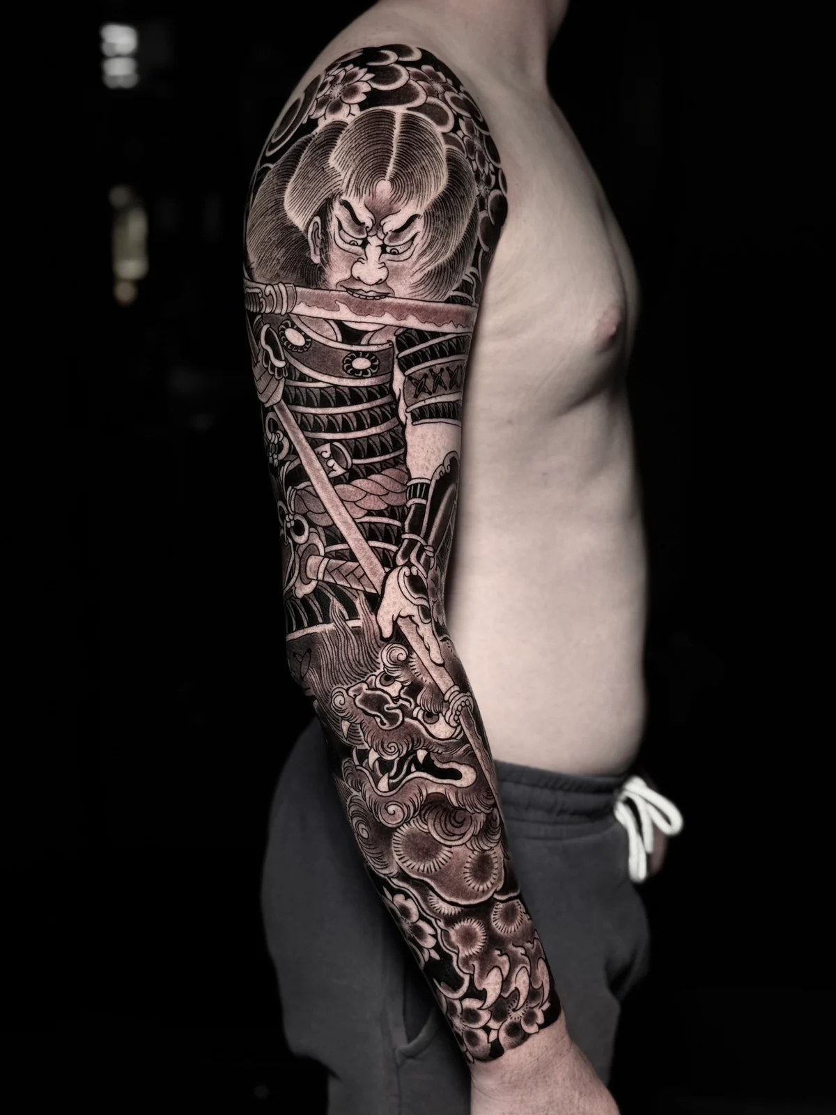 Japanese black and grey full sleeve with samurai biting a katana blade and snarling shishi amid cherry blossoms and clouds at Bloodline Tattoo Phuket.