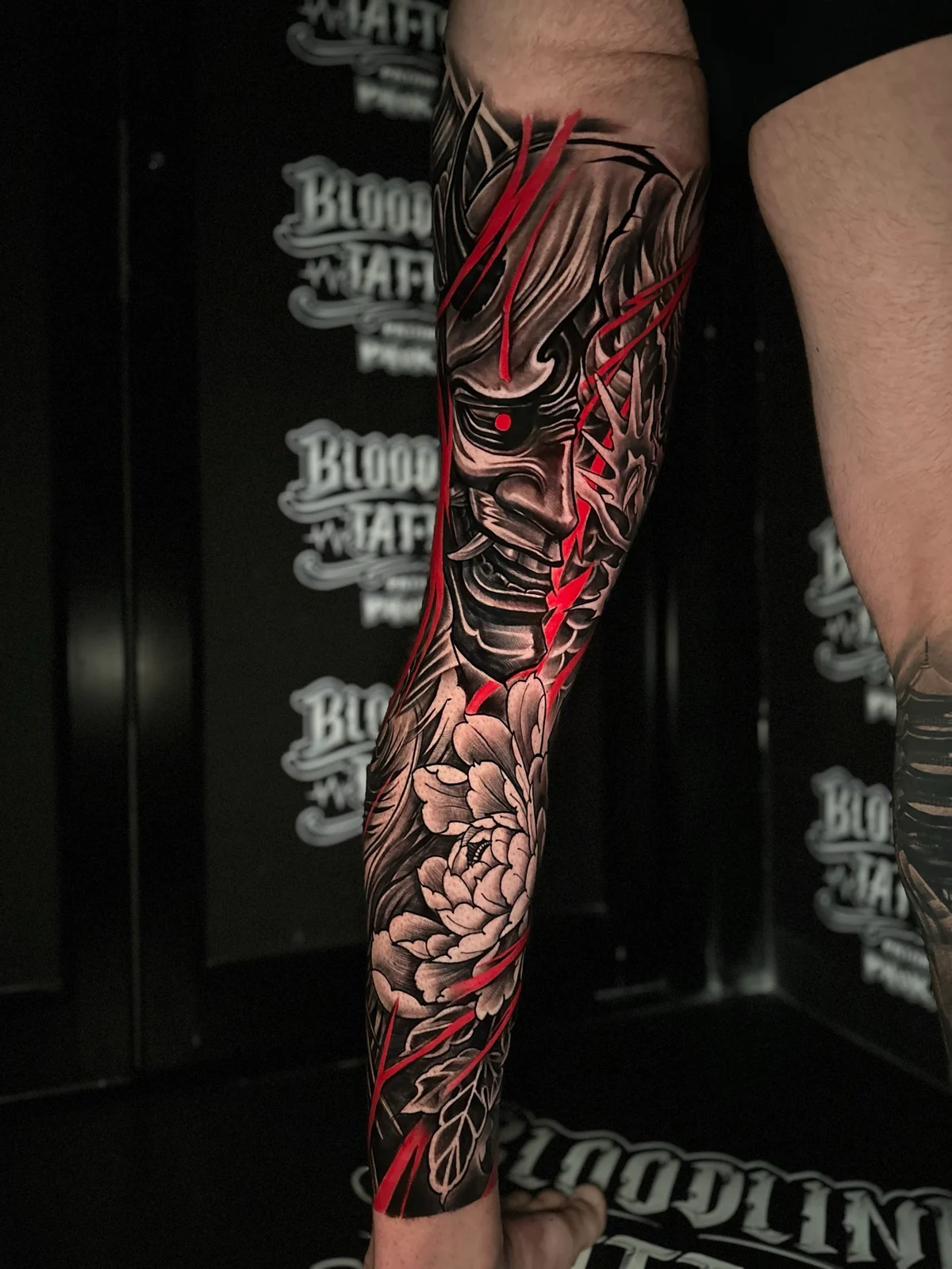 Japanese lower leg tattoo with Hannya mask, red eye accent, blooming peony, and jagged red lightning slashes at Bloodline Tattoo Phuket.