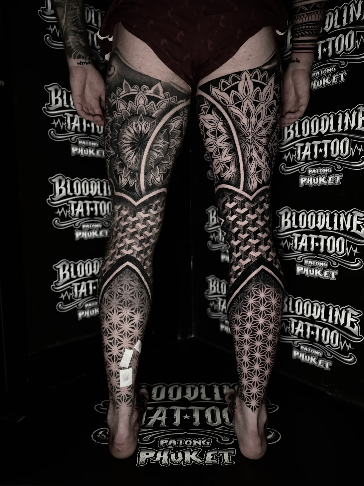 Posterior dual full leg mandala work with upper-thigh lotus mandalas, isometric 3D cube blackwork on the calves, and Flower of Life dotwork near the ankles, at Bloodline Tattoo Phuket.