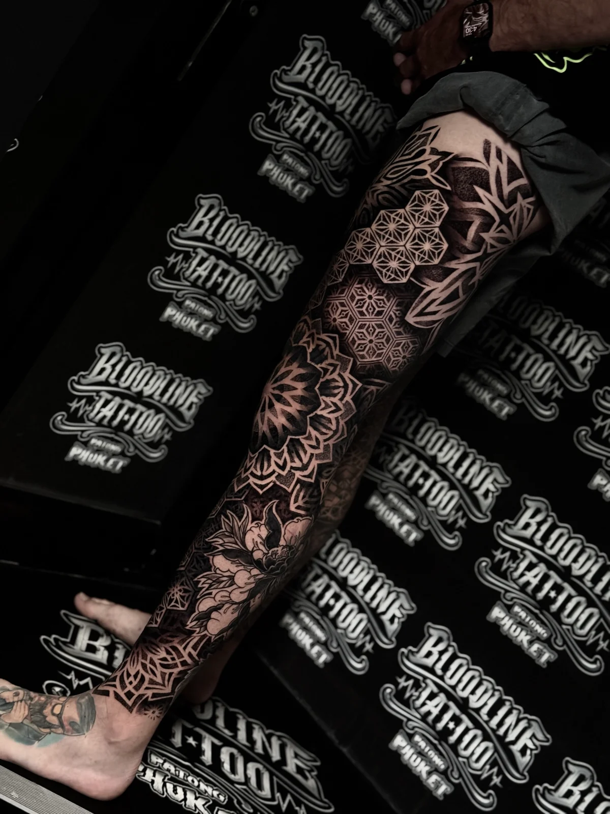 Full leg mandala sleeve centered on a knee lotus mandala with honeycomb thigh grids and shaded peony florals wrapping the lower shin, completed at Bloodline Tattoo Phuket.
