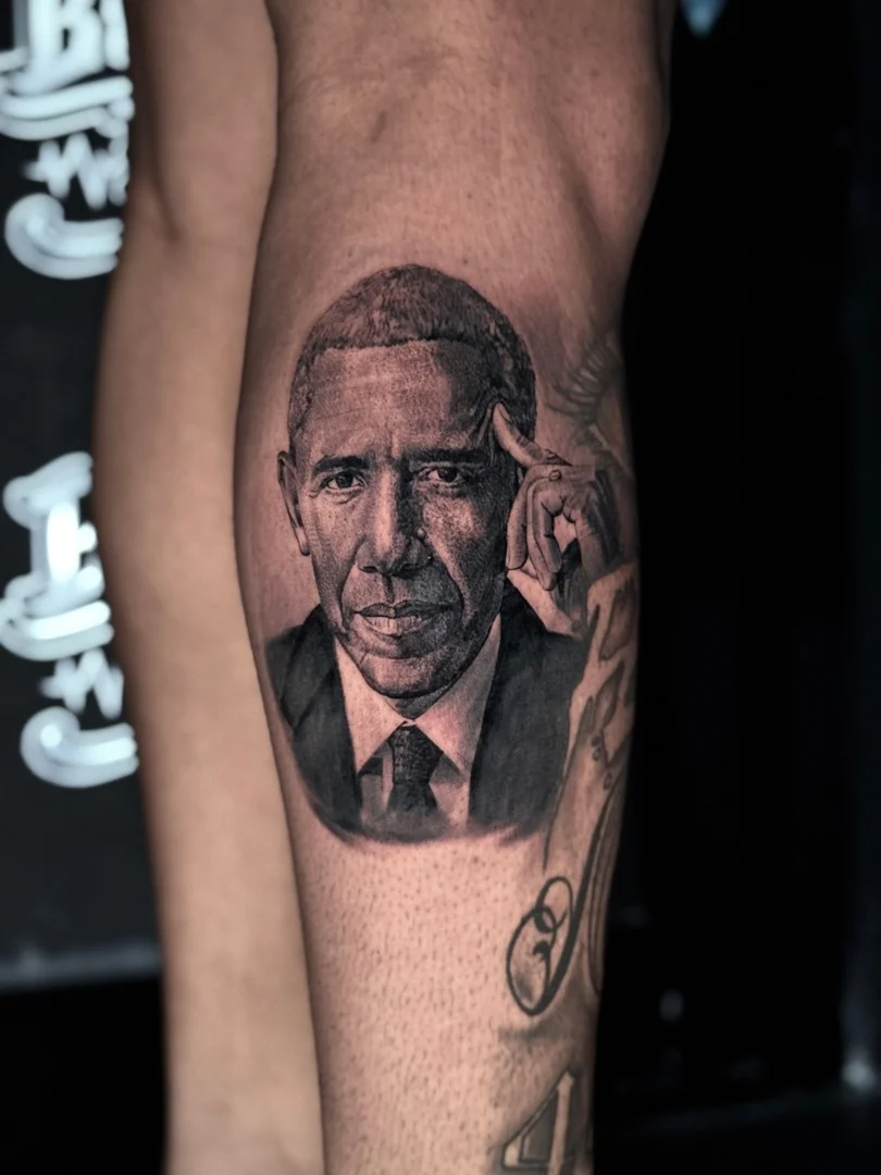 Black and grey portrait realism forearm tattoo of Barack Obama in a suit with a thoughtful hand-to-temple pose at Bloodline Tattoo Phuket.