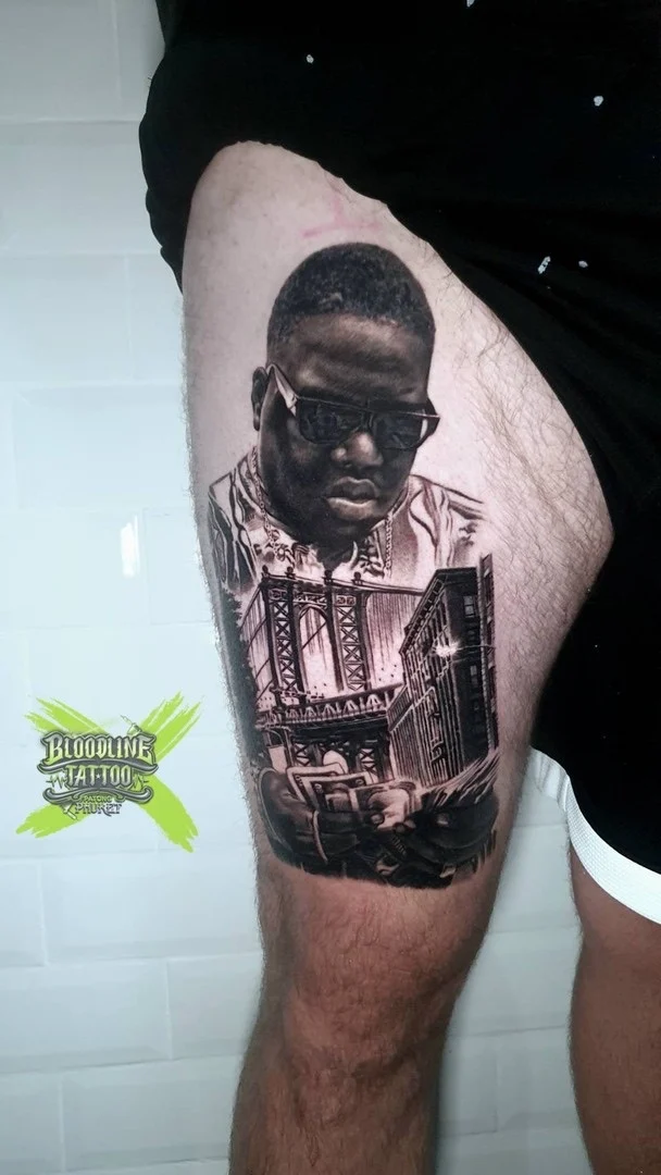 Black and grey portrait realism thigh tattoo of The Notorious B.I.G. with NYC bridge skyline and cash hands at Bloodline Tattoo Phuket.