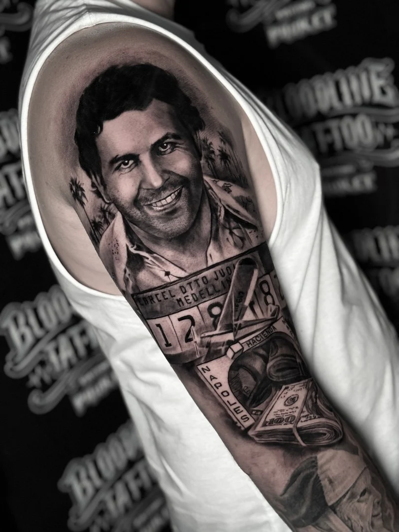 Black and grey portrait realism shoulder tattoo of Pablo Escobar with Medellin prison placard, plane, and Hacienda Napoles stack at Bloodline Tattoo Phuket.