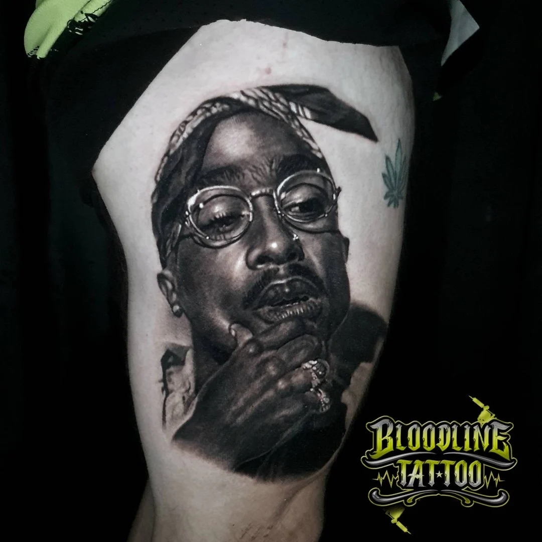 Black and grey portrait realism thigh tattoo of Tupac Shakur in bandana and round glasses at Bloodline Tattoo Phuket.