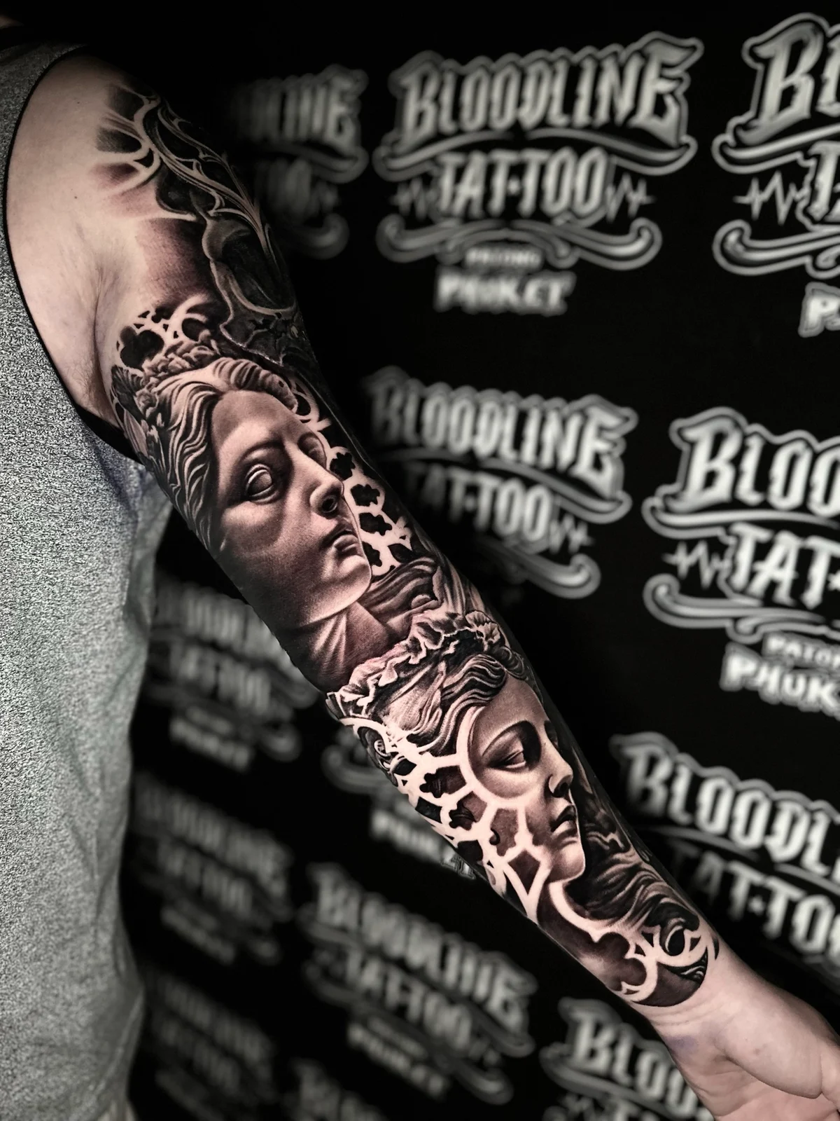 Detailed black and grey realism sleeve tattoo featuring two classical female sculpture faces surrounded by intricate architectural patterns at Bloodline Tattoo Phuket.