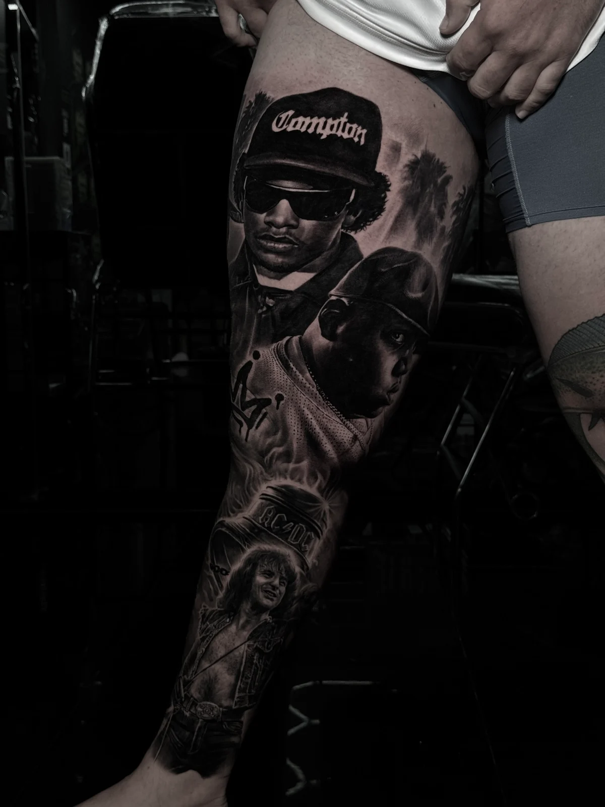 Detailed black and grey realism leg sleeve tattoo featuring portraits of Eazy-E, The Notorious B.I.G., and Angus Young from AC/DC at Bloodline Tattoo Phuket.