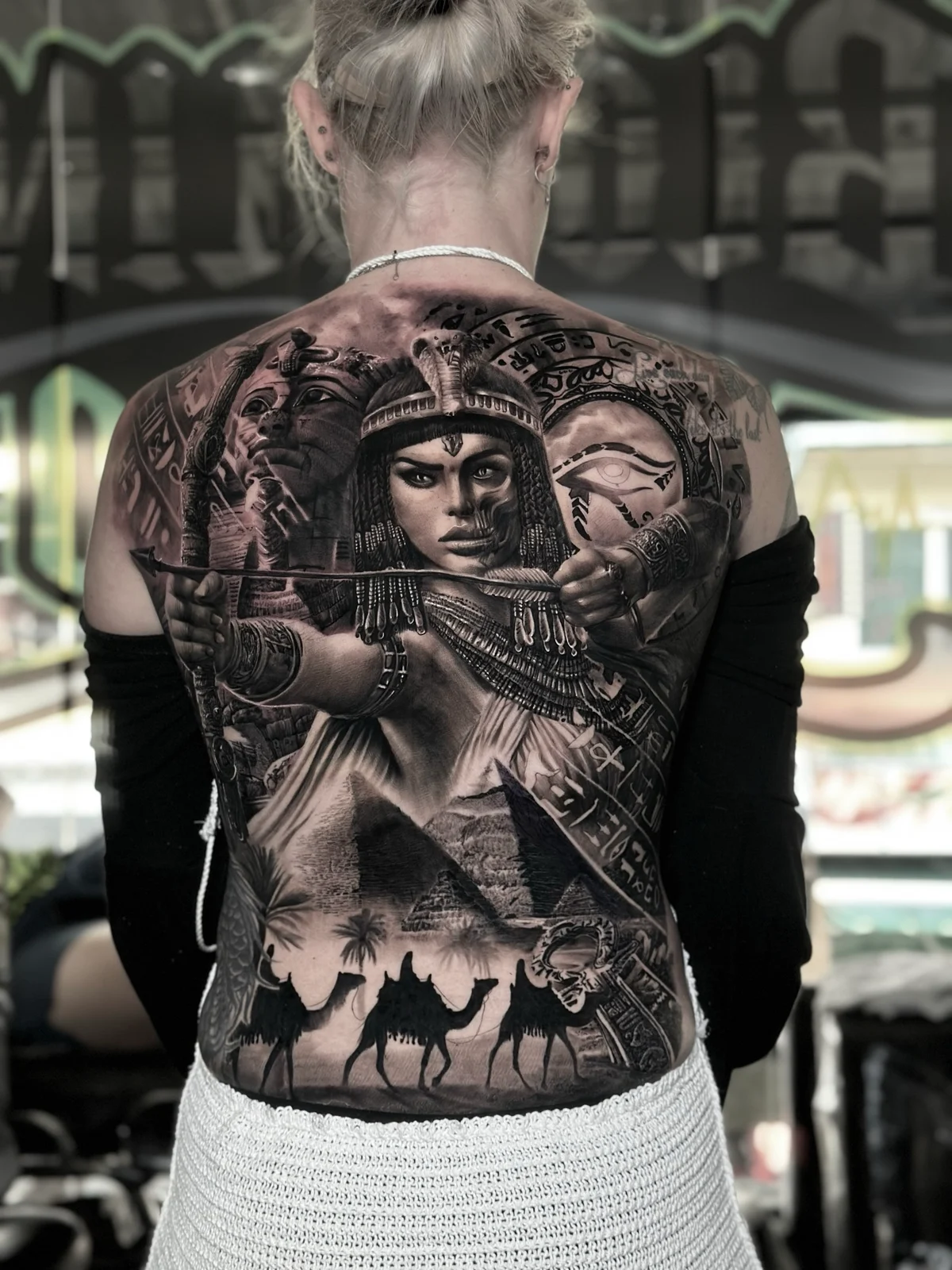 Detailed black and grey realism full back tattoo featuring an Egyptian queen with bow, Pharaoh statue, Eye of Horus, Pyramids of Giza, and a camel caravan at Bloodline Tattoo Phuket.