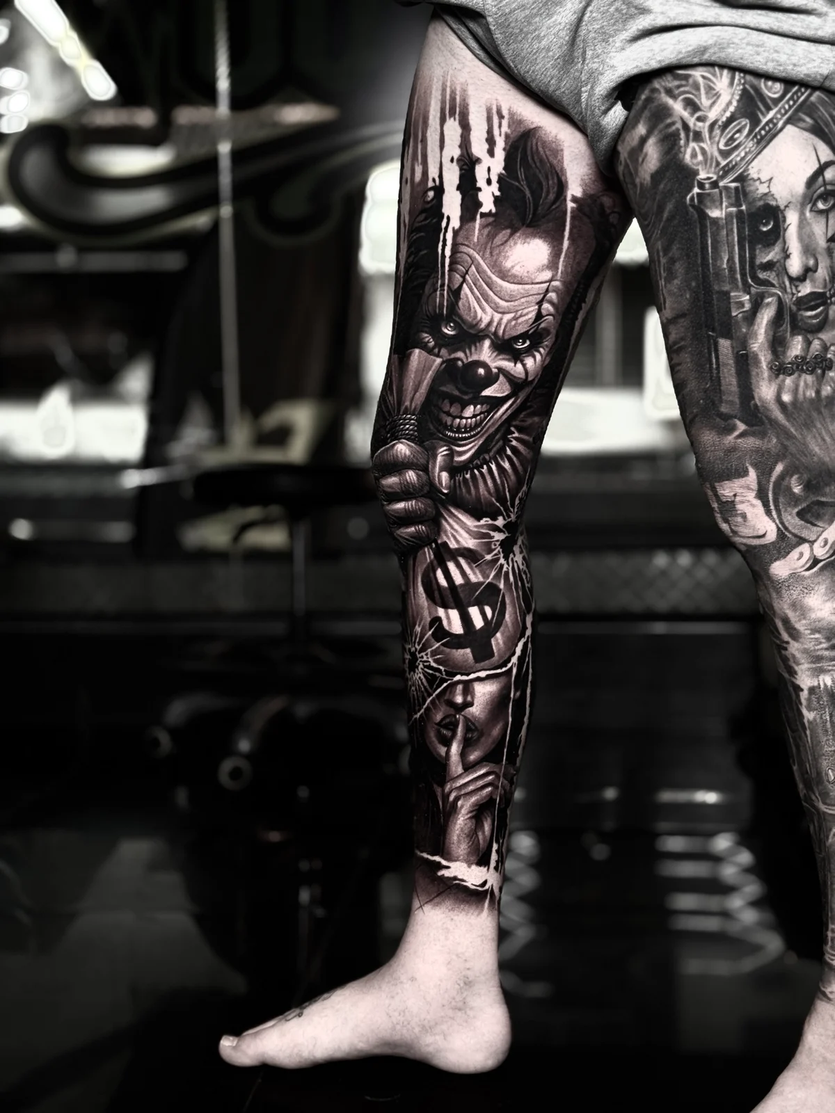 Detailed black and grey realism leg sleeve tattoo featuring a menacing horror clown holding a money bag and a woman making a shushing gesture at Bloodline Tattoo Phuket.