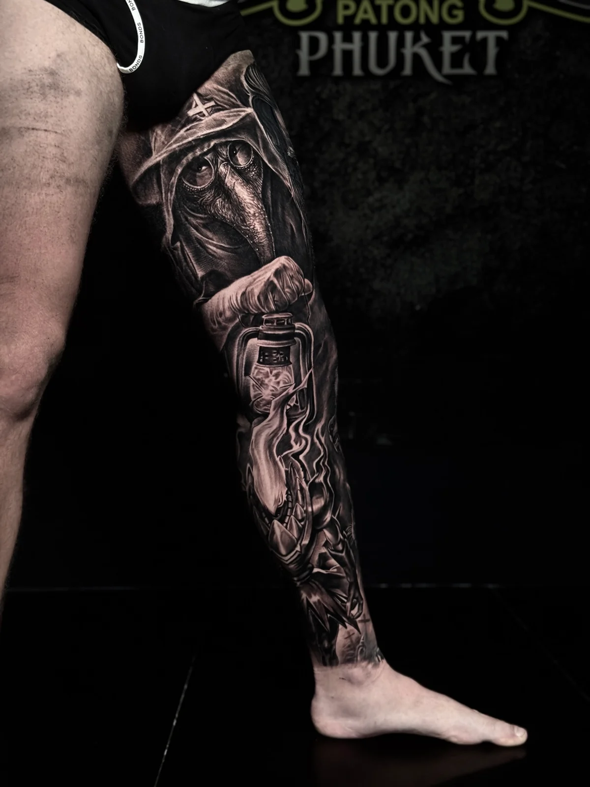 Detailed black and grey realism full leg sleeve tattoo featuring a plague doctor, glowing lantern with smoke, demonic face, and graveyard crosses at Bloodline Tattoo Phuket.